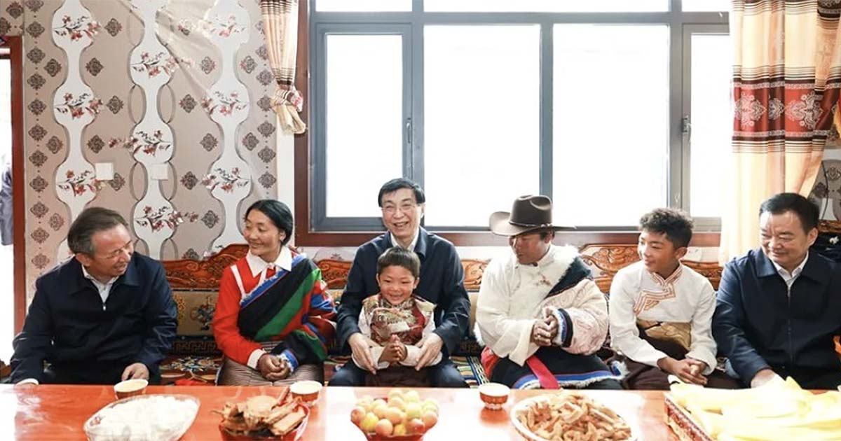 Wang Huning zit lachend tussen een Tibetaanse familie in traditionele kleding tijdens een geënsceneerd huisbezoek in Tsamdha, met een Tibetaans kind op schoot