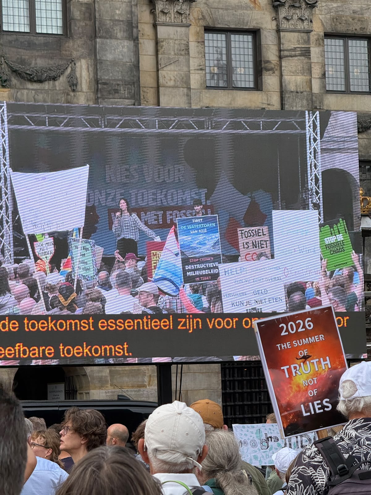 Videoscherm op de Dam met een spreker op het podium; tussen de protestborden in beeld het bord "Tibet, de watertoren van Azië — stop China's destructieve milieubeleid in Tibet"