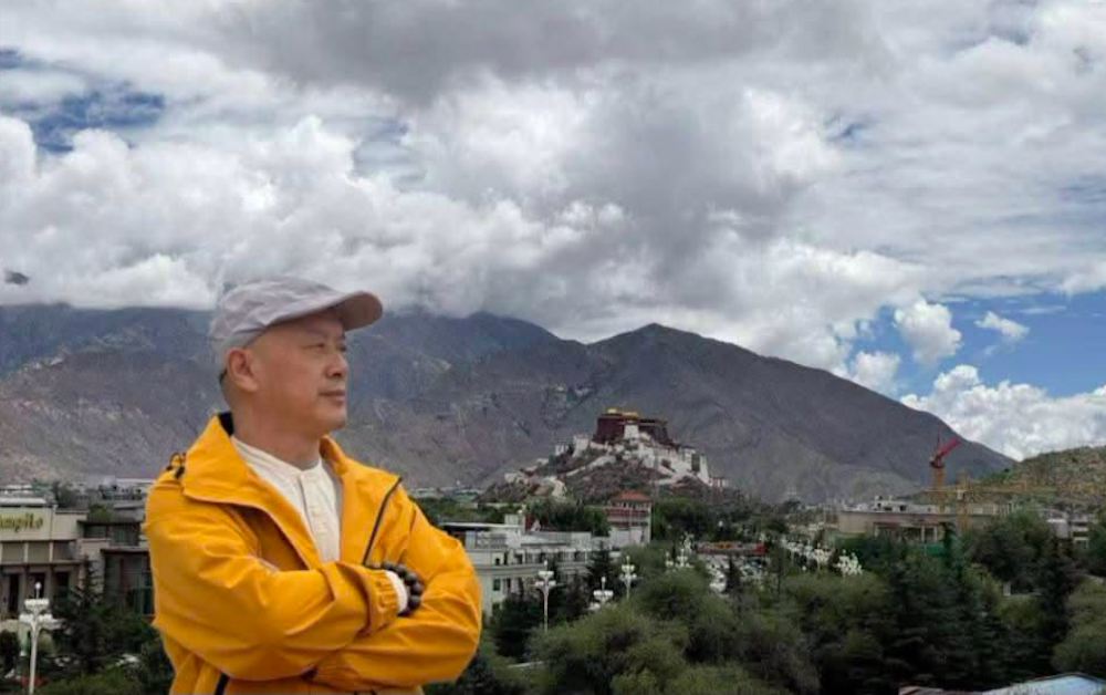 Zhang Yi in een gele jas en met pet, armen over elkaar, met op de achtergrond het Potala-paleis in Lhasa onder een wolkenlucht